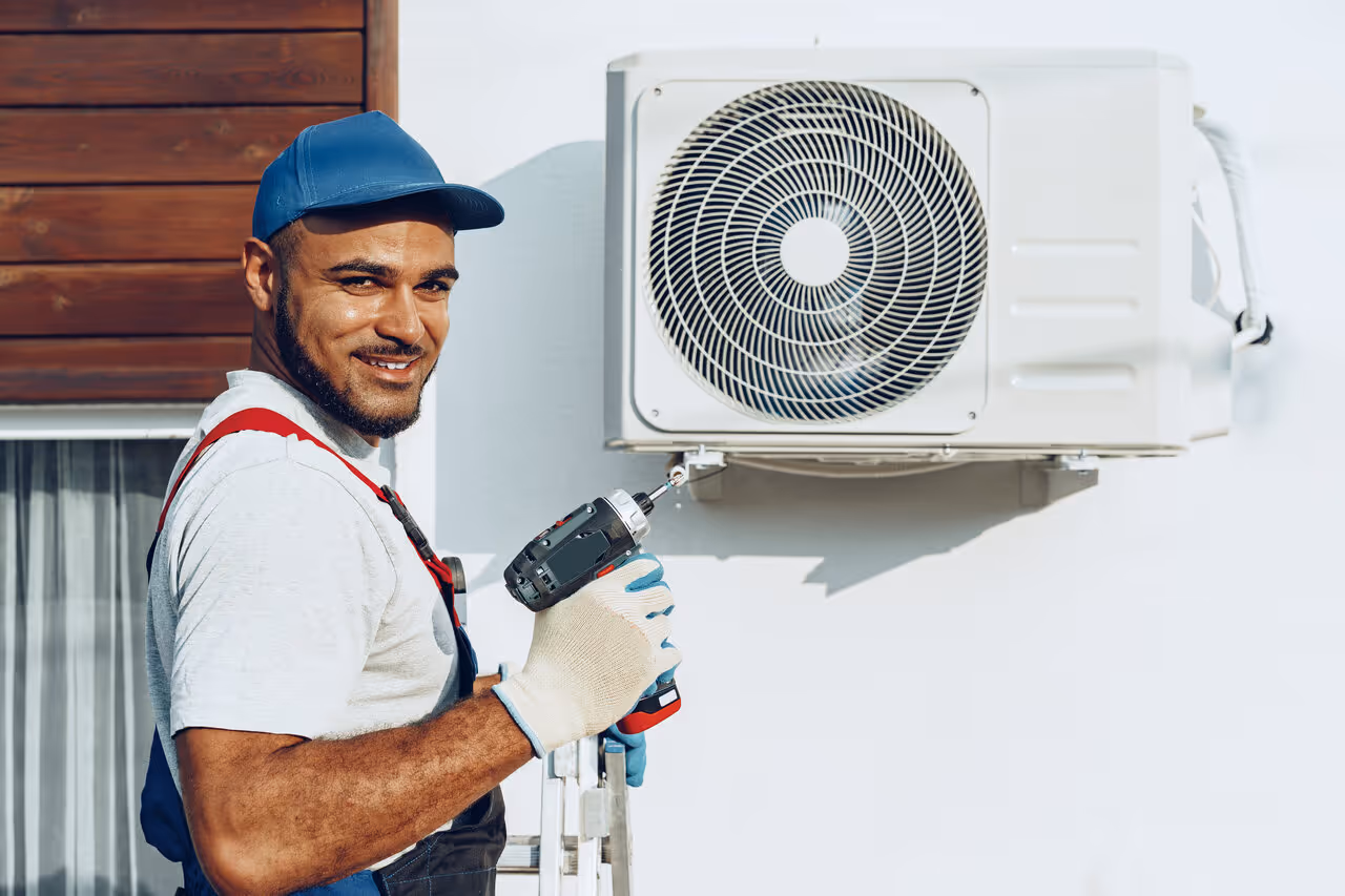 A smiling technician in a blue cap uses a power drill to install an air conditioning unit on a white wall