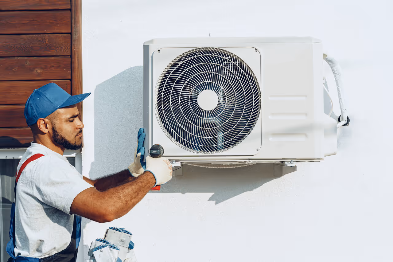 A technician in a blue cap and gloves installs an air conditioning unit on a white wall. 