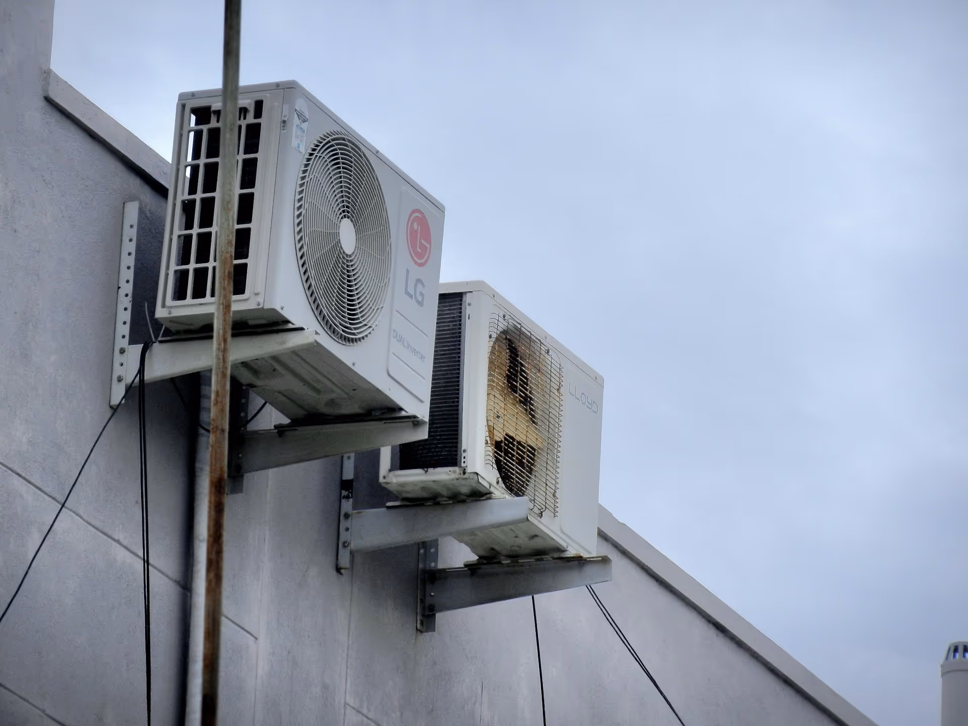 Two air conditioning units mounted on an exterior wall under a cloudy sky.