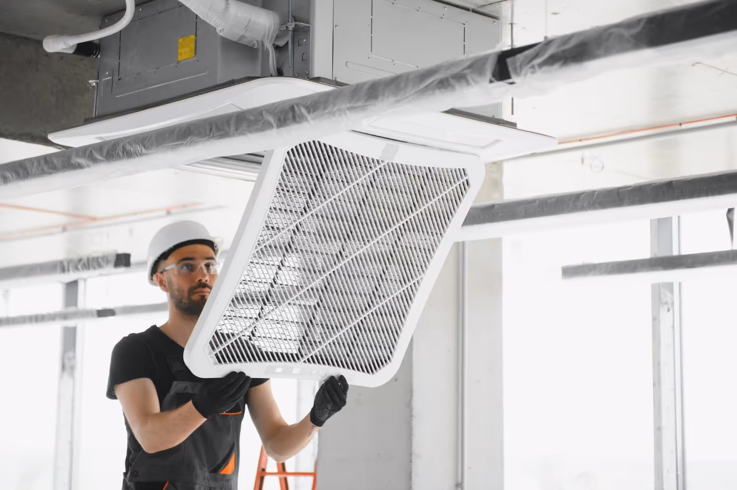 A worker in a hard hat and gloves installs a ventilation cover on a ceiling-mounted HVAC unit in a well-lit