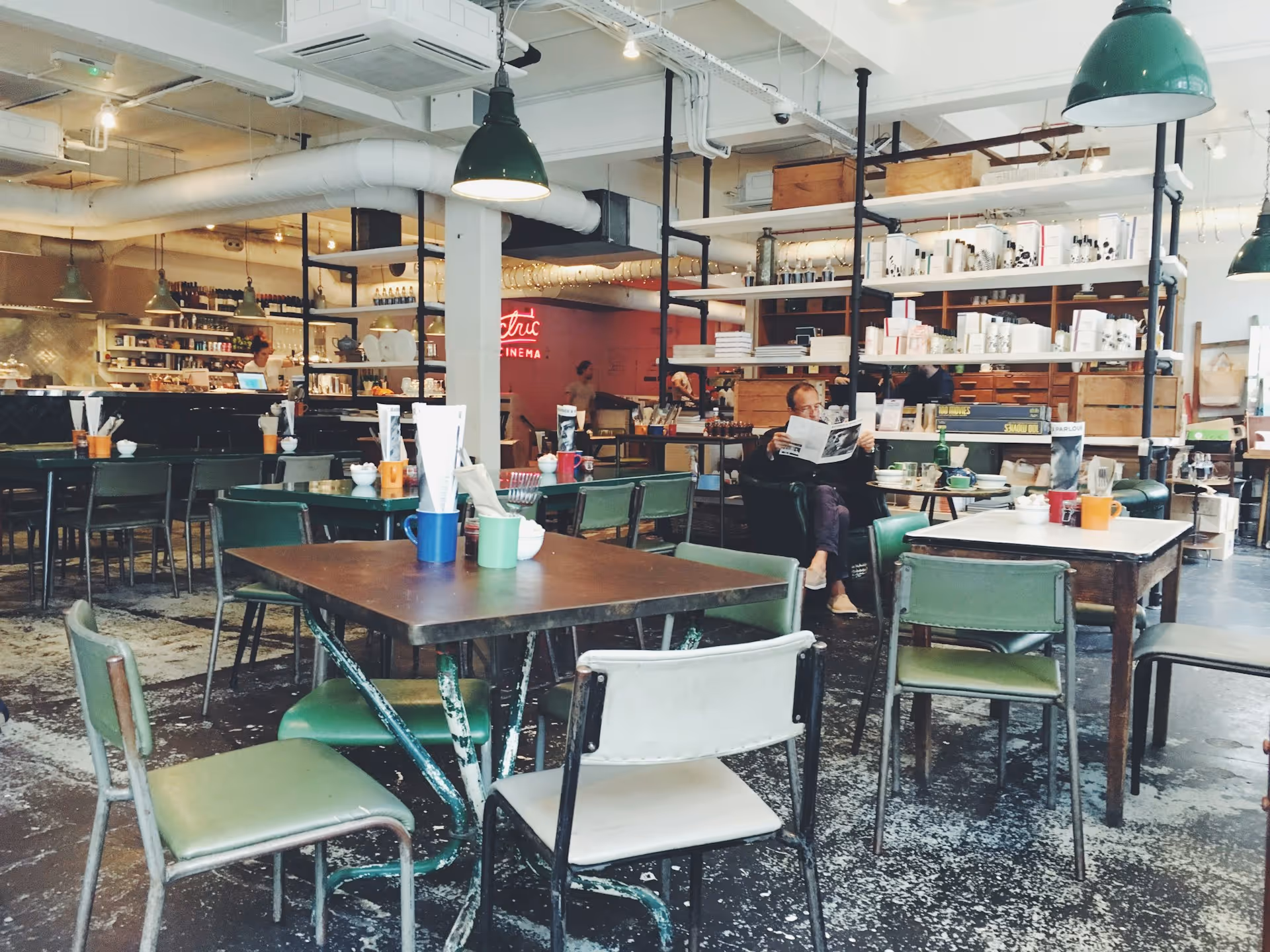 Cozy cafe interior with rustic decor, green chairs, wooden tables, and industrial lighting. 