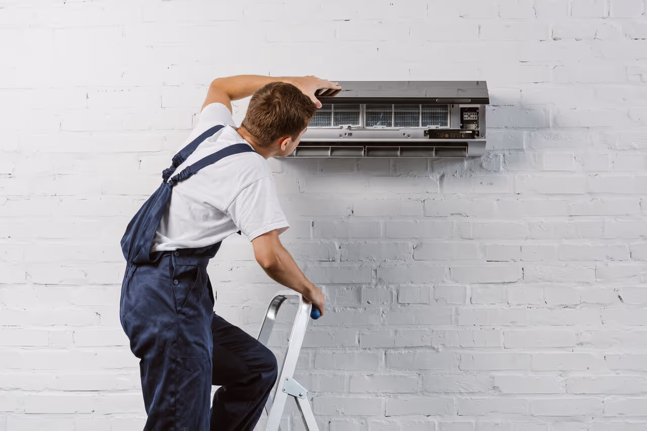 A technician wearing overalls stands on a ladder fixing an air conditioner on a white brick wall