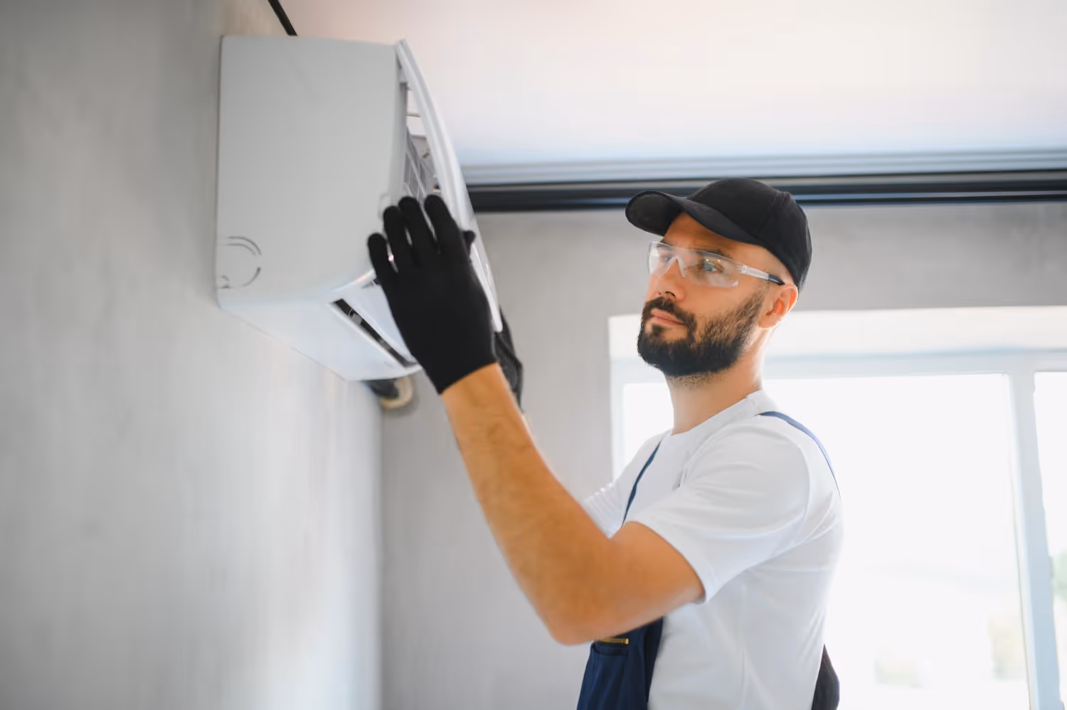 A man wearing safety goggles and gloves inspects an air conditioning unit on a wall,
