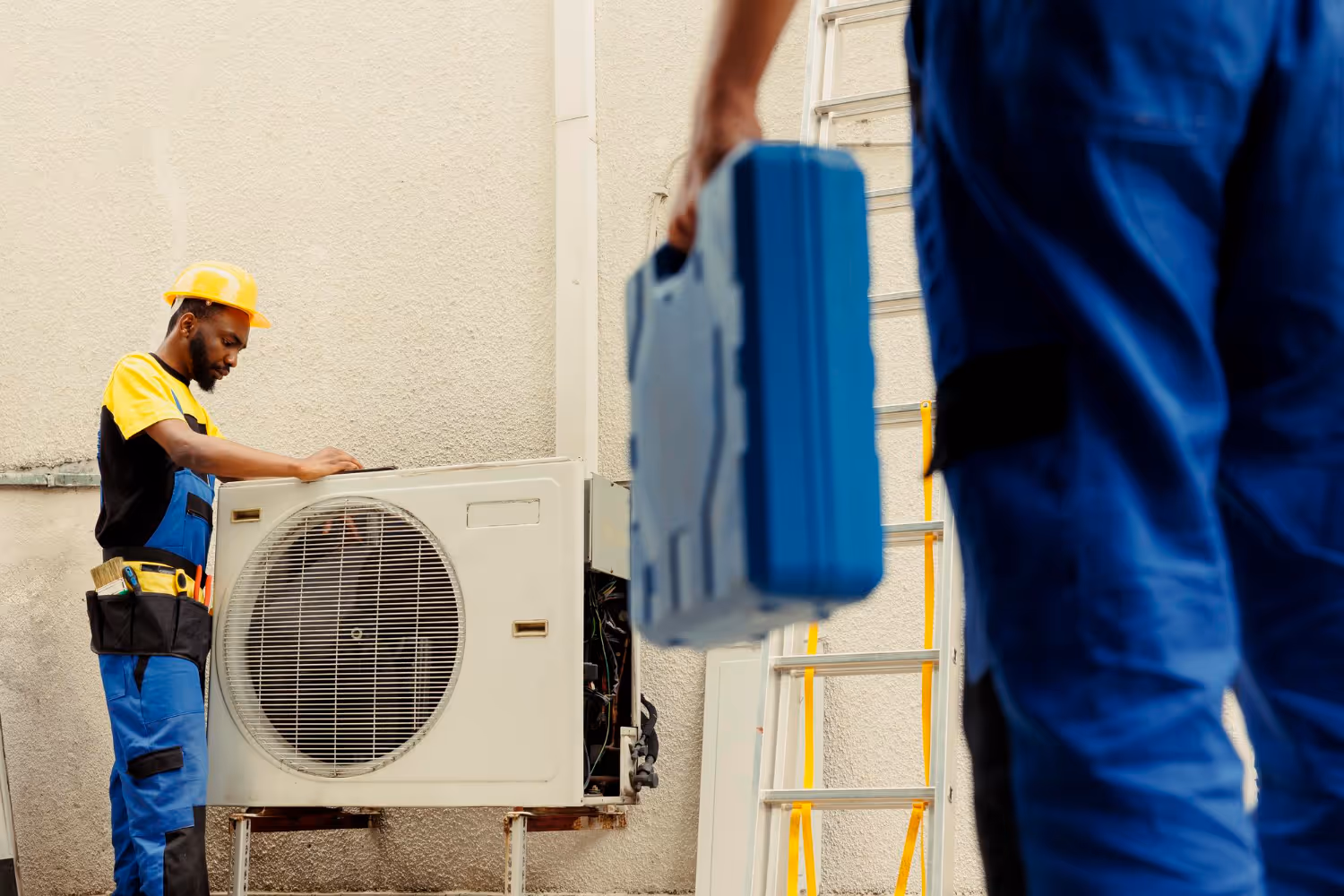 A technician in a blue uniform and yellow hard hat repairs an outdoor air conditioning unit.