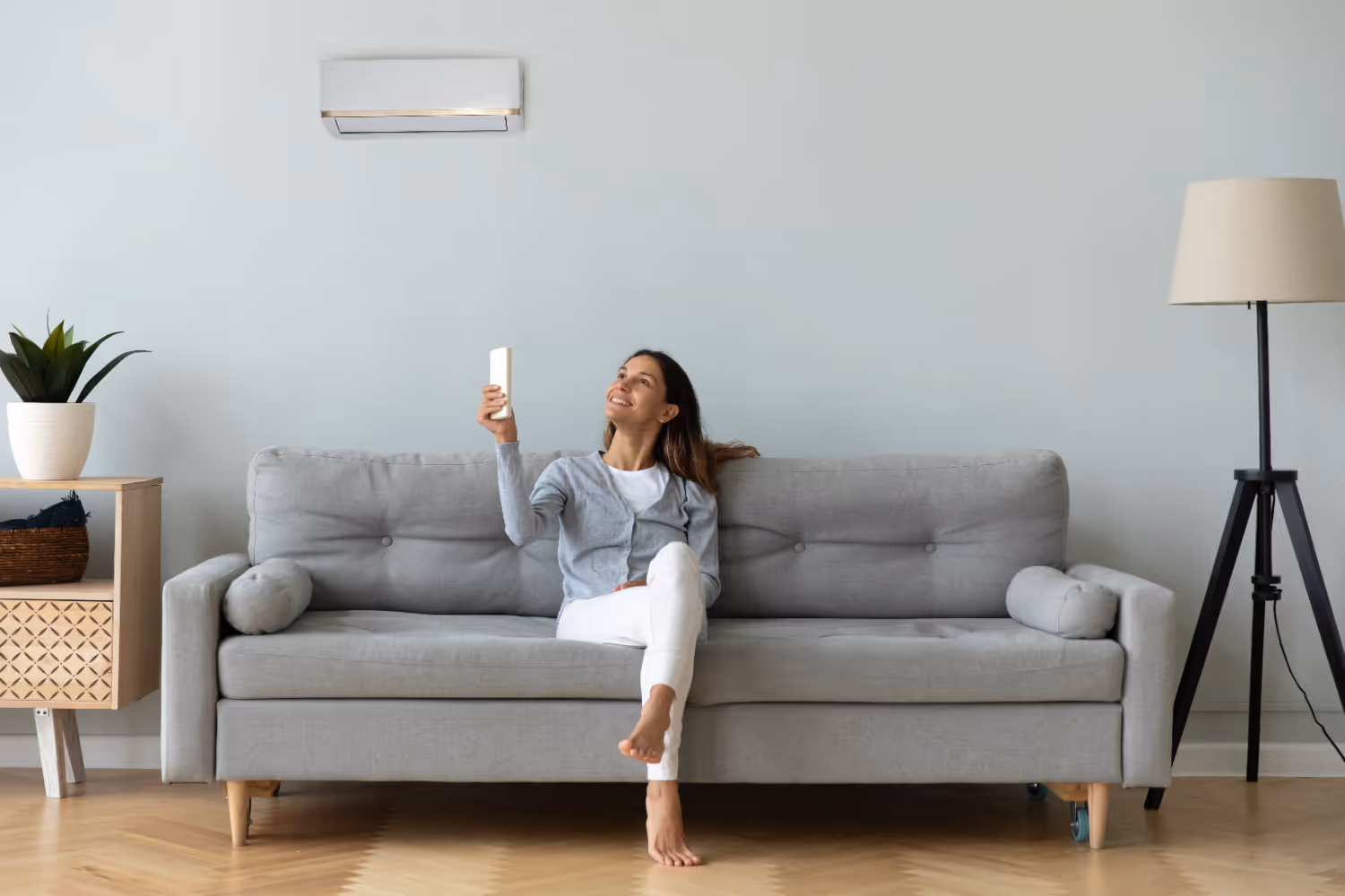 A woman sits on a gray sofa, smiling as she points a remote at an air conditioner. 