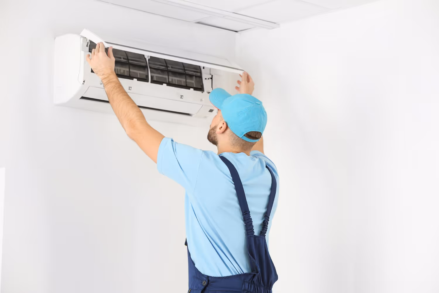 A technician in blue uniform and cap is servicing a wall-mounted air conditioner in a bright room.