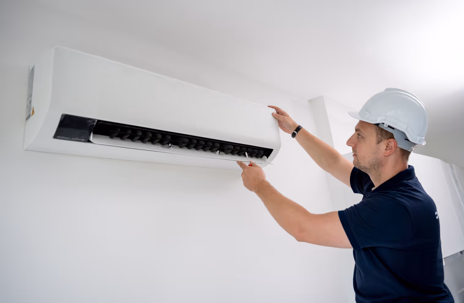 A technician in a hard hat and blue shirt inspects a white wall-mounted air conditioning unit.