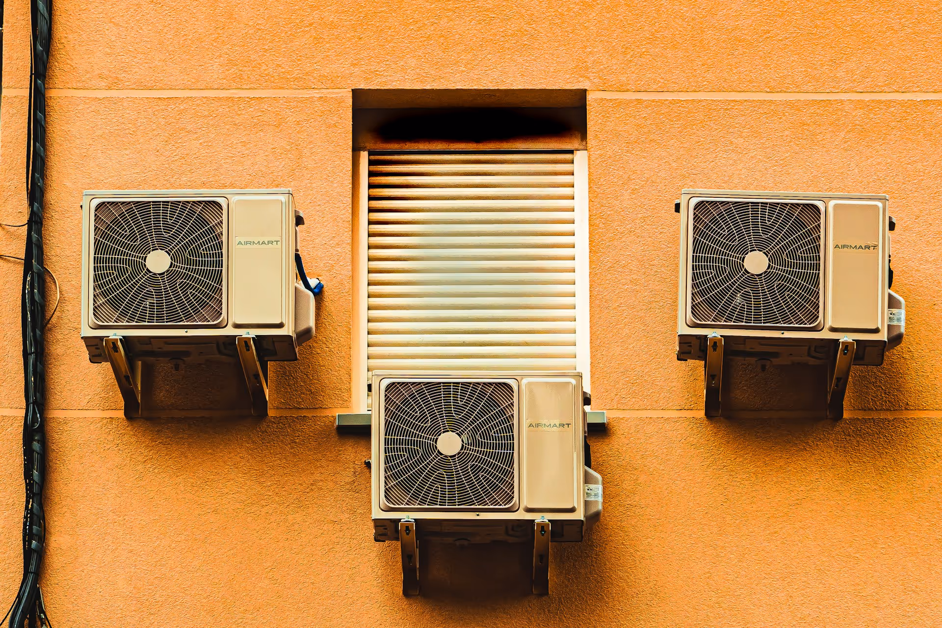 Three air conditioning units are mounted on an orange stucco wall around a closed, louvered vent.