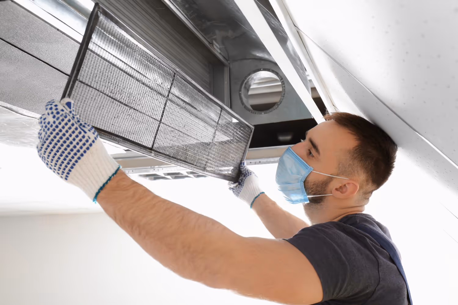 A person wearing gloves and a face mask installs a filter into a ceiling air vent. 