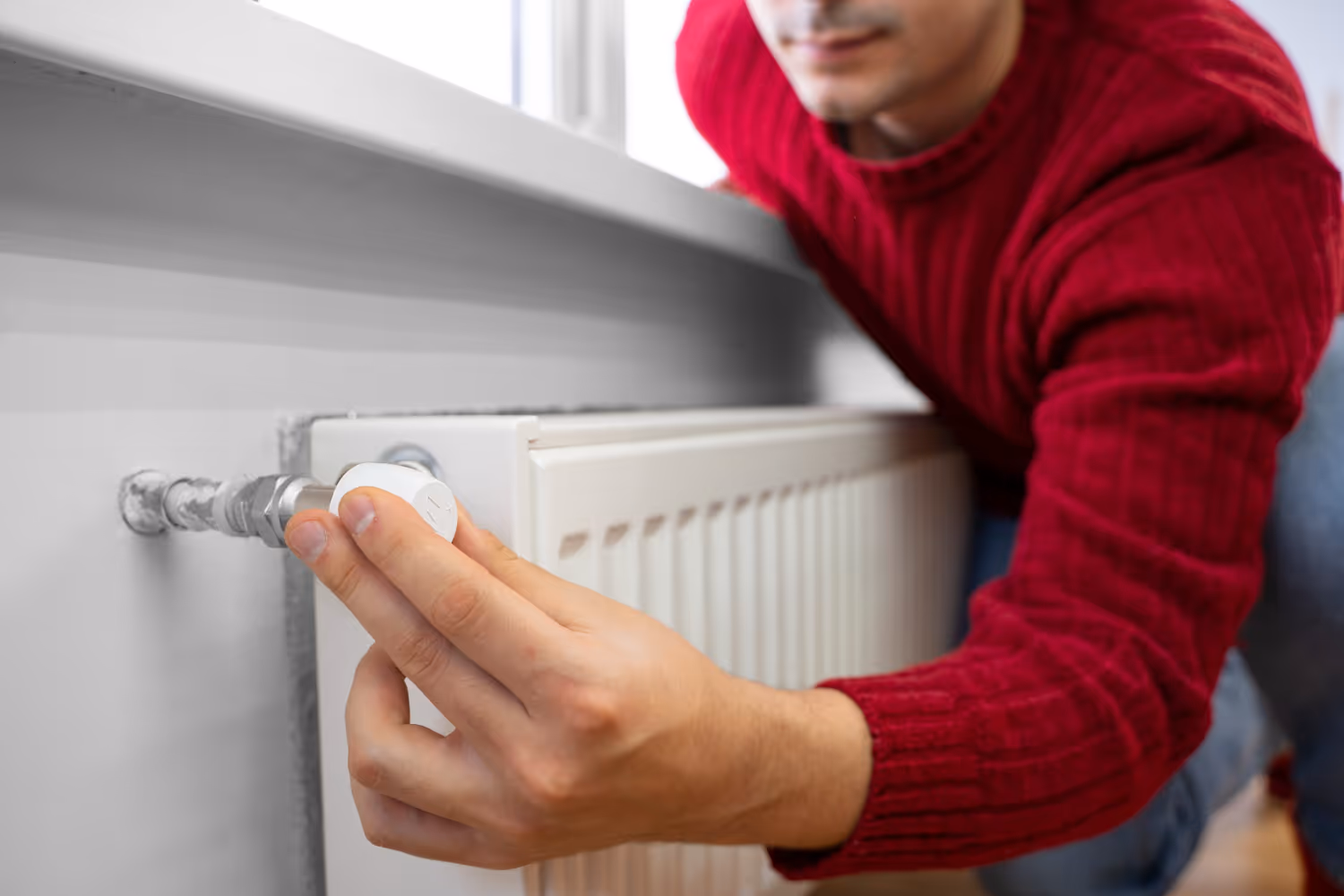 A person in a red sweater adjusts a thermostat valve on a white radiator under a window