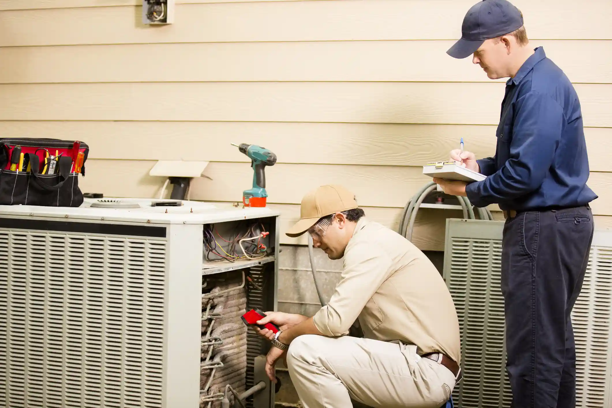 Two HVAC technicians are servicing an outdoor air conditioning or heat pump condenser unit next to a tan-colored wall. One technician, kneeling and wearing a khaki shirt and a tan cap, is inspecting the internal coils and components with a red handheld tool. The second technician, standing and wearing a dark blue uniform and cap, is holding a clipboard and taking notes. A tool bag and a cordless drill are visible on the top of the unit.