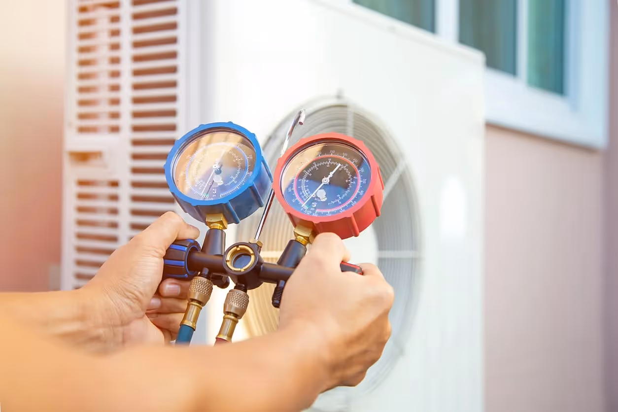 A close-up of a person's hands holding a manifold gauge set with red and blue dials, about to connect them to a heat pump unit.