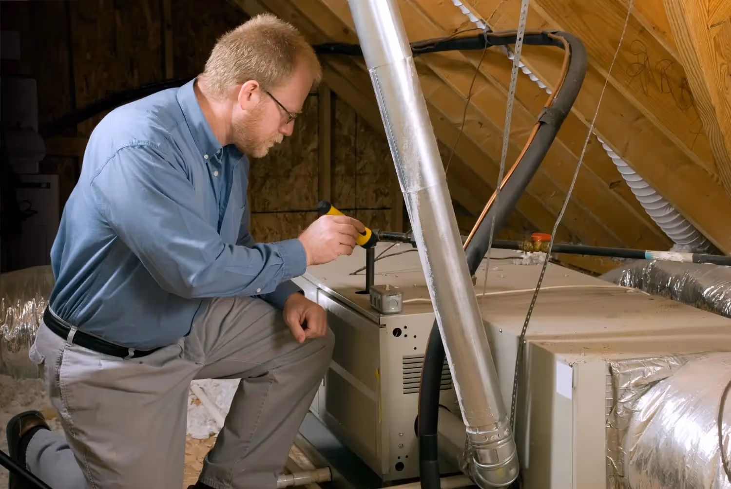 A bespectacled male HVAC technician wearing a blue shirt and khakis kneels in a dusty attic, using a small flashlight to inspect the internal connections of a horizontal air handler or furnace unit.