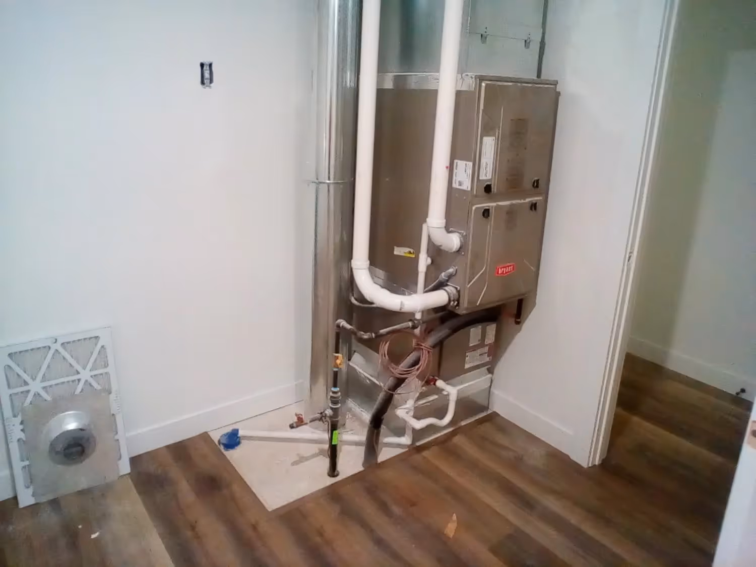 A recently installed HVAC air handler or furnace is mounted vertically in a utility closet with white walls and wood-look flooring. The unit is connected to large metal ductwork and white PVC venting pipes, with a new air filter lying on the floor nearby.