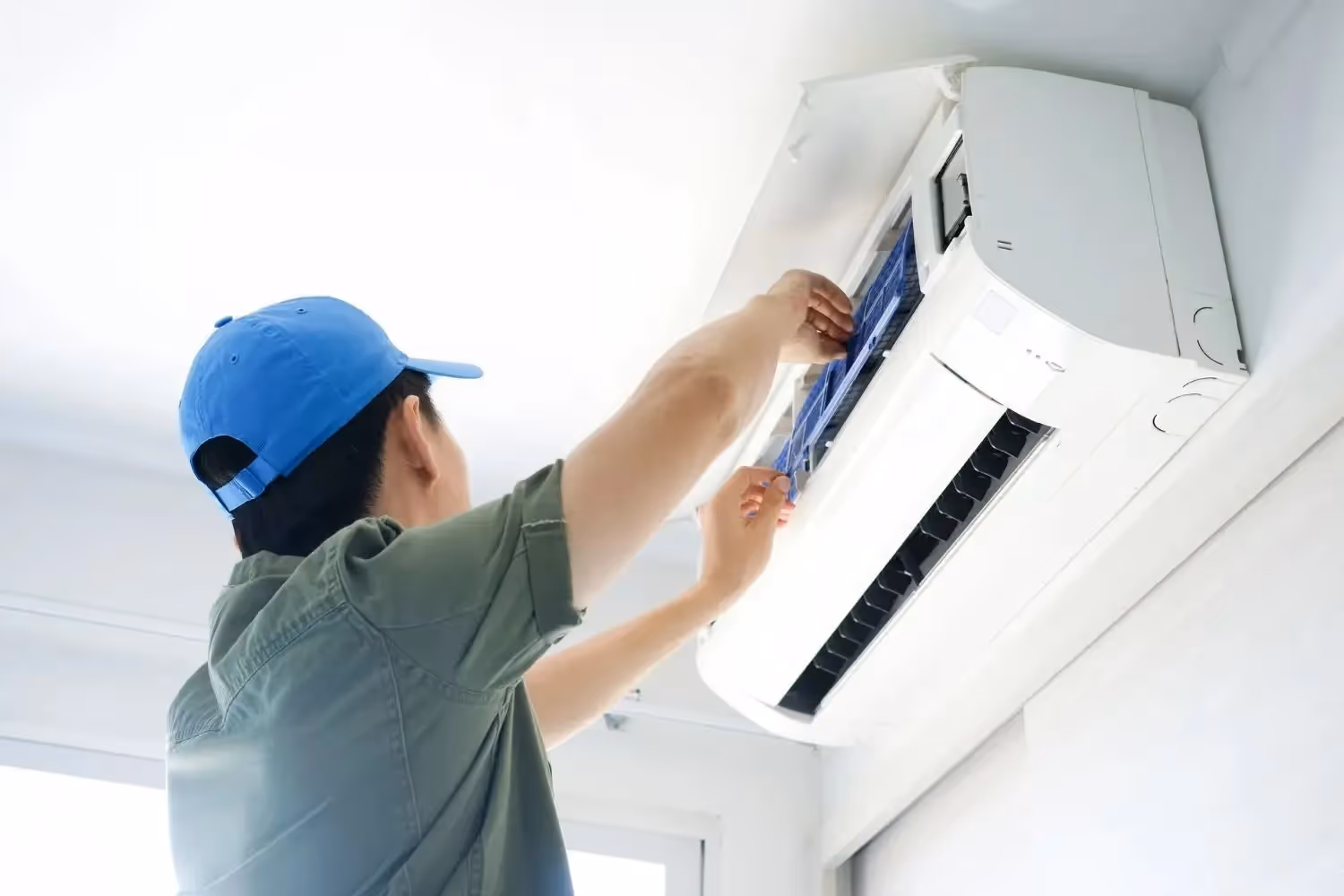 An HVAC technician wearing a blue hat is cleaning the filter of a wall-mounted ductless air conditioner.