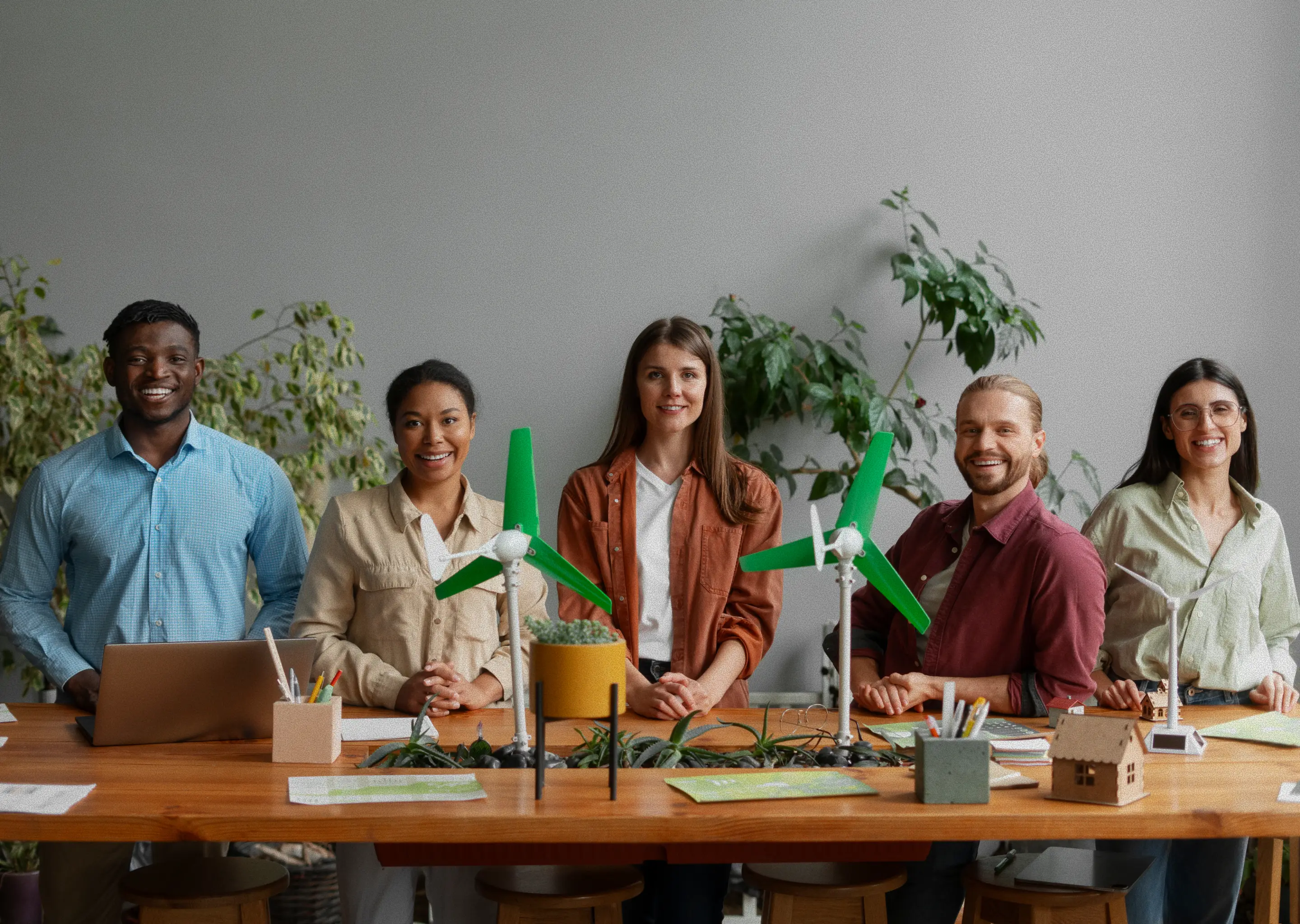 A full-length, eye-level shot shows a diverse group of five smiling colleagues posing behind a long wooden table. The group consists of two men and three women.