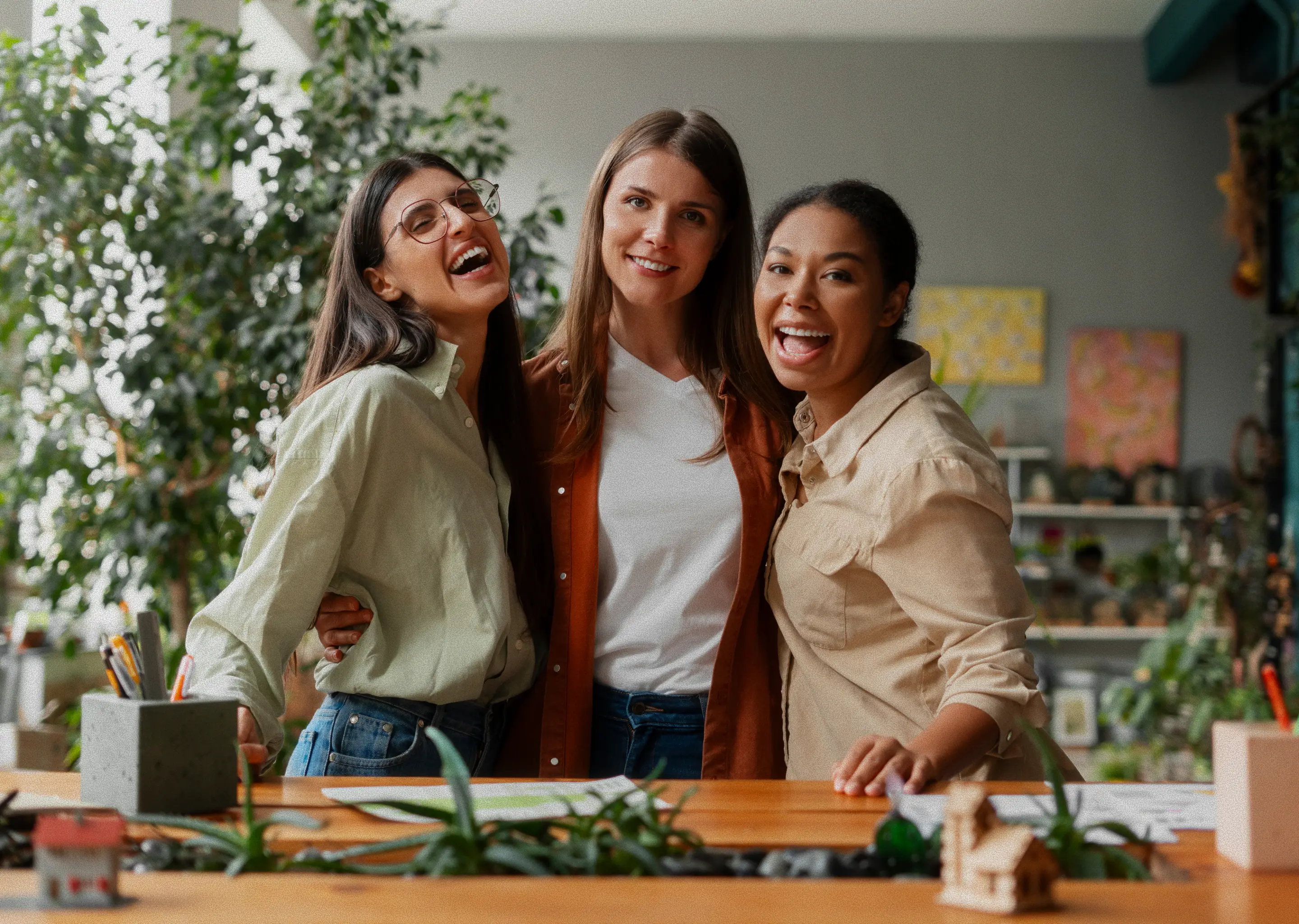 A close-up, eye-level shot shows three women of different ethnicities smiling and laughing while standing behind a desk.