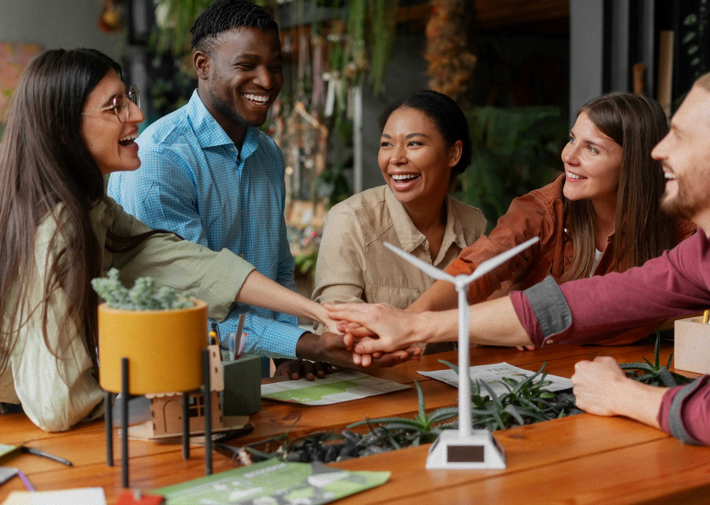 A diverse group of five smiling colleagues, three women and two men, are gathered around a wooden table in an office, stacking their hands on top of each other in a show of teamwork.