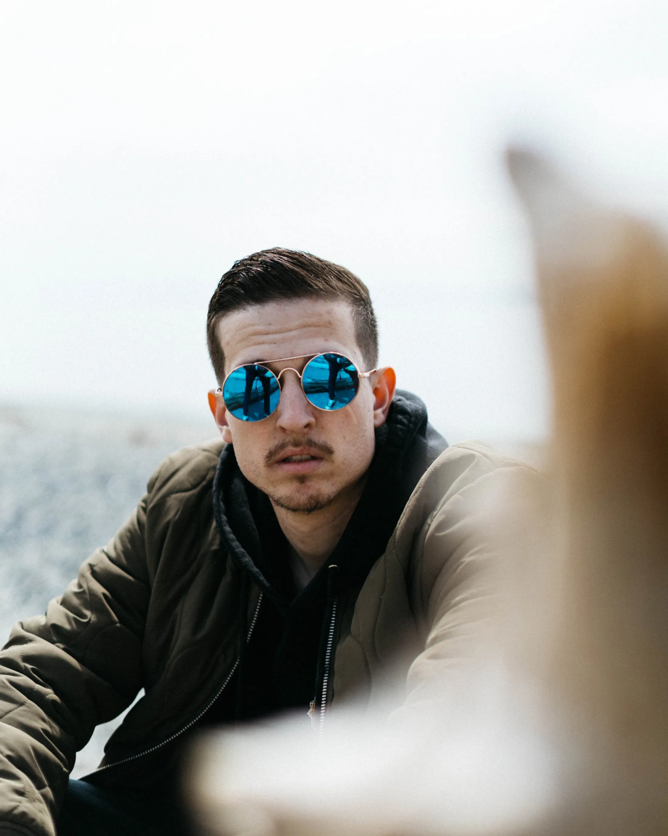 Person in blue reflective sunglasses and olive jacket looking serious