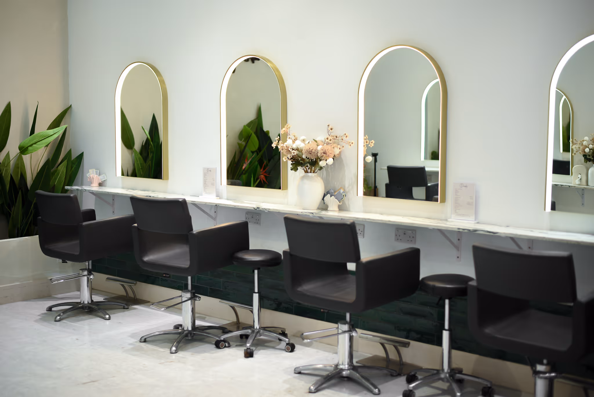 Modern hair salon station with four black chairs, illuminated arched mirrors, and decorative plants and flowers.