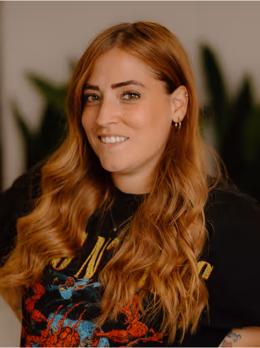 Smiling woman with long wavy red hair wearing a black graphic t-shirt standing in front of blurred green plants.