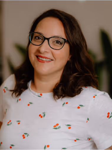 Smiling woman with dark curly hair wearing black glasses and a white shirt with red cherry patterns.