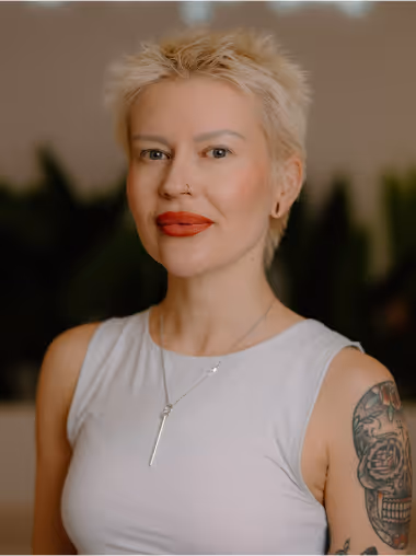 Portrait of a woman with short blonde hair, red lipstick, a nose ring, and a tattoo on her upper arm, wearing a white sleeveless top and a silver necklace.