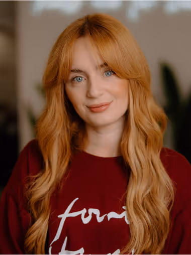 Woman with long wavy red hair and blue eyes wearing a burgundy shirt with white script text, smiling softly.