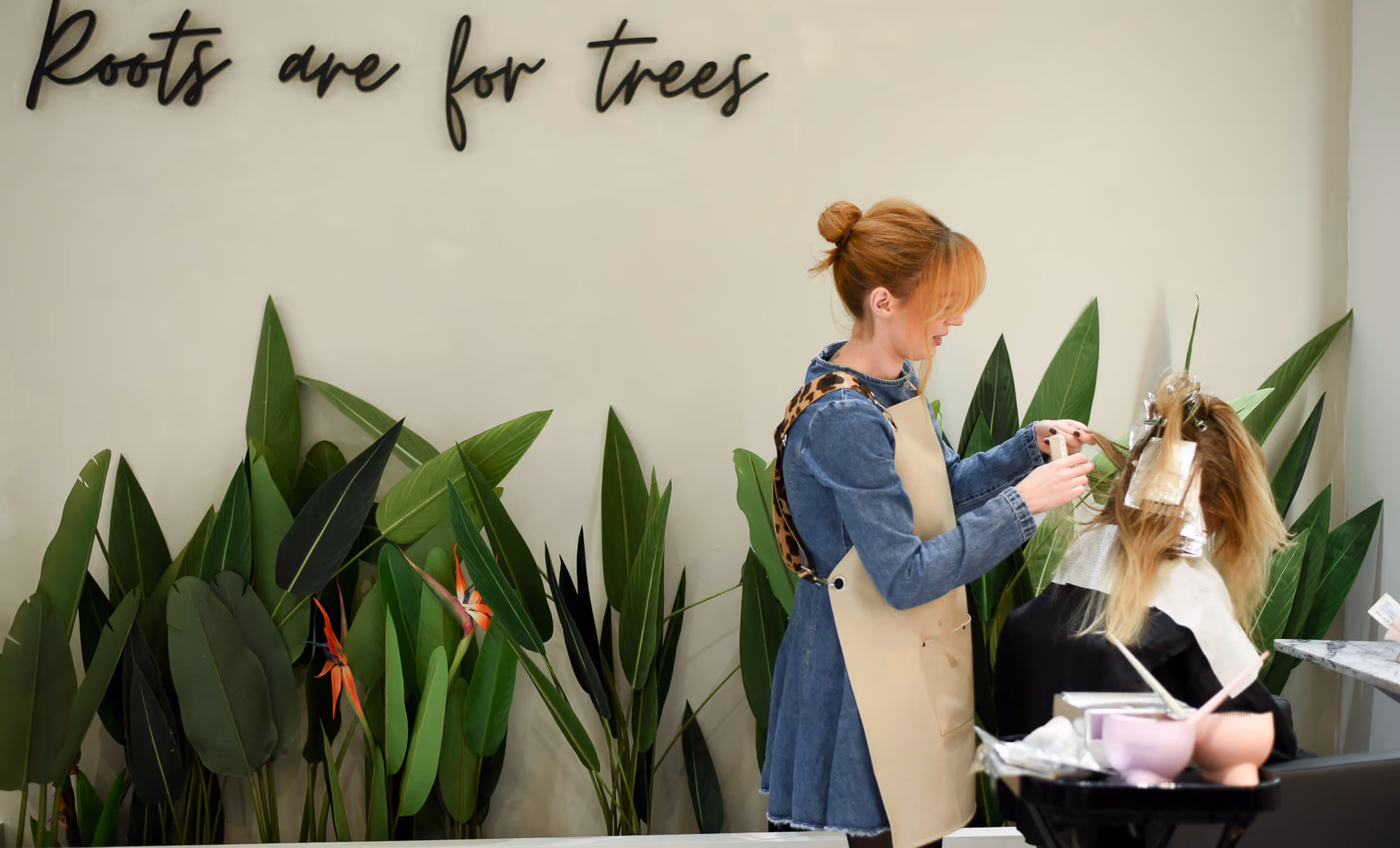 A stylist in a denim dress with a cream apron applying foils to a client. In the background is a wall with a sign that says 'Roots are for trees' and a row of large real tropical plants below.