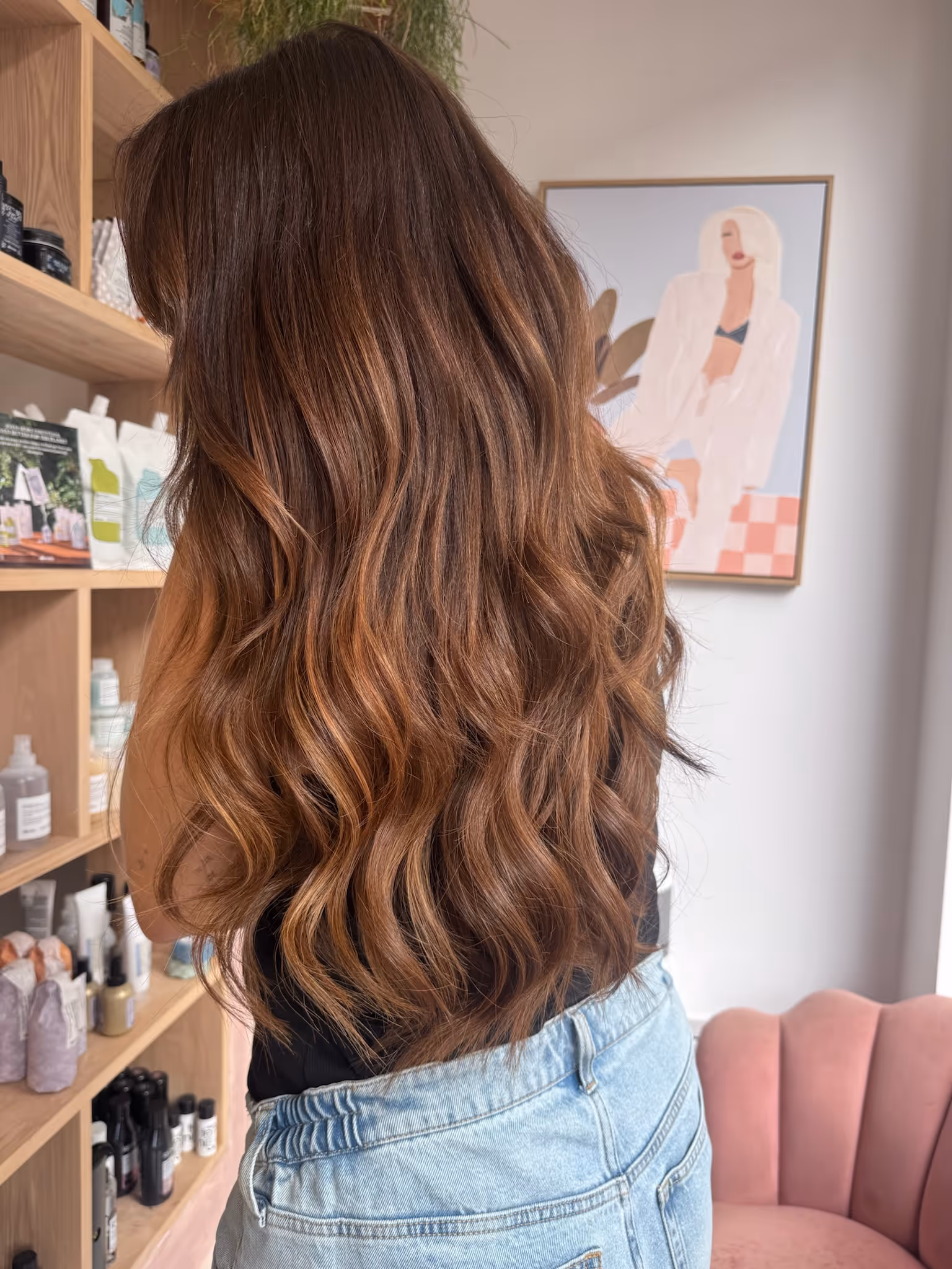 Woman with long wavy brown hair with caramel highlights standing in front of wooden shelves with beauty products.