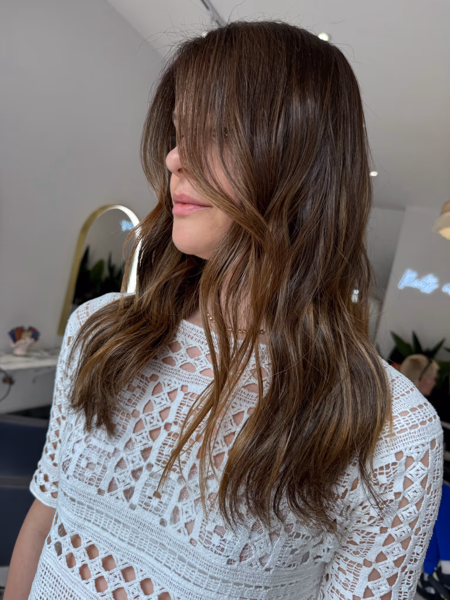 Woman with long layered brown hair styled in loose waves, wearing a white crocheted top.