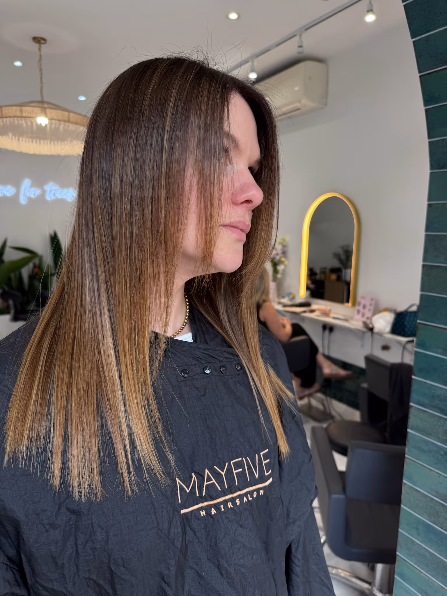 Side view of a woman with straight, shoulder-length brown hair with subtle highlights in a hair salon.