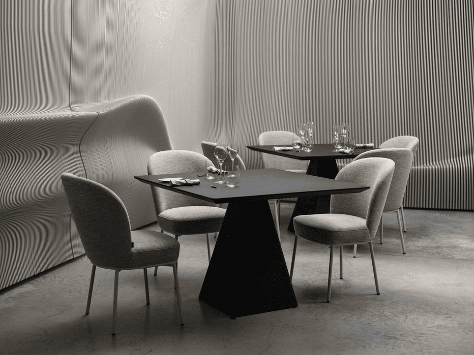 Modern restaurant interior with black square tables and grey upholstered chairs against a curved, ribbed wall design.