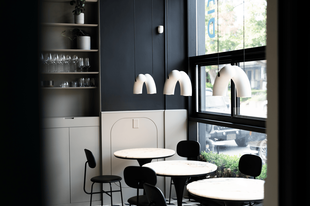 Modern café interior with round marble tables, black chairs, white pendant lights, and large window showing parked cars outside.