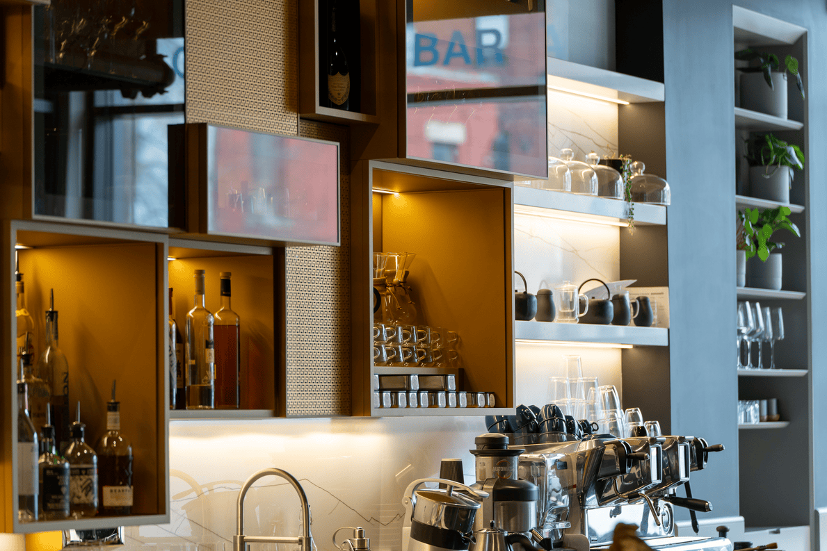 Modern bar area with shelves holding liquor bottles, glassware, and coffee equipment under warm lighting.