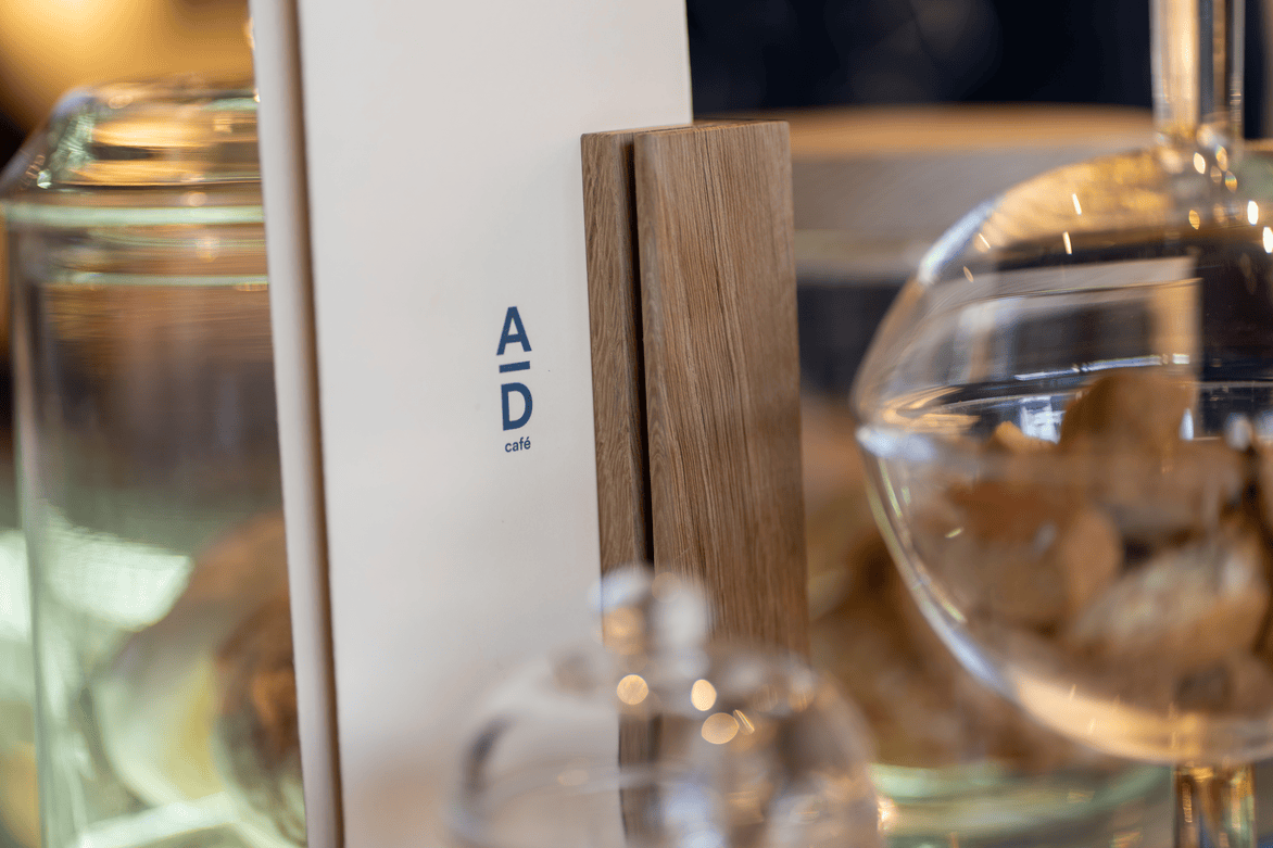Close-up of a café menu with the logo 'A over D café' held by a wooden stand, surrounded by glass display jars.