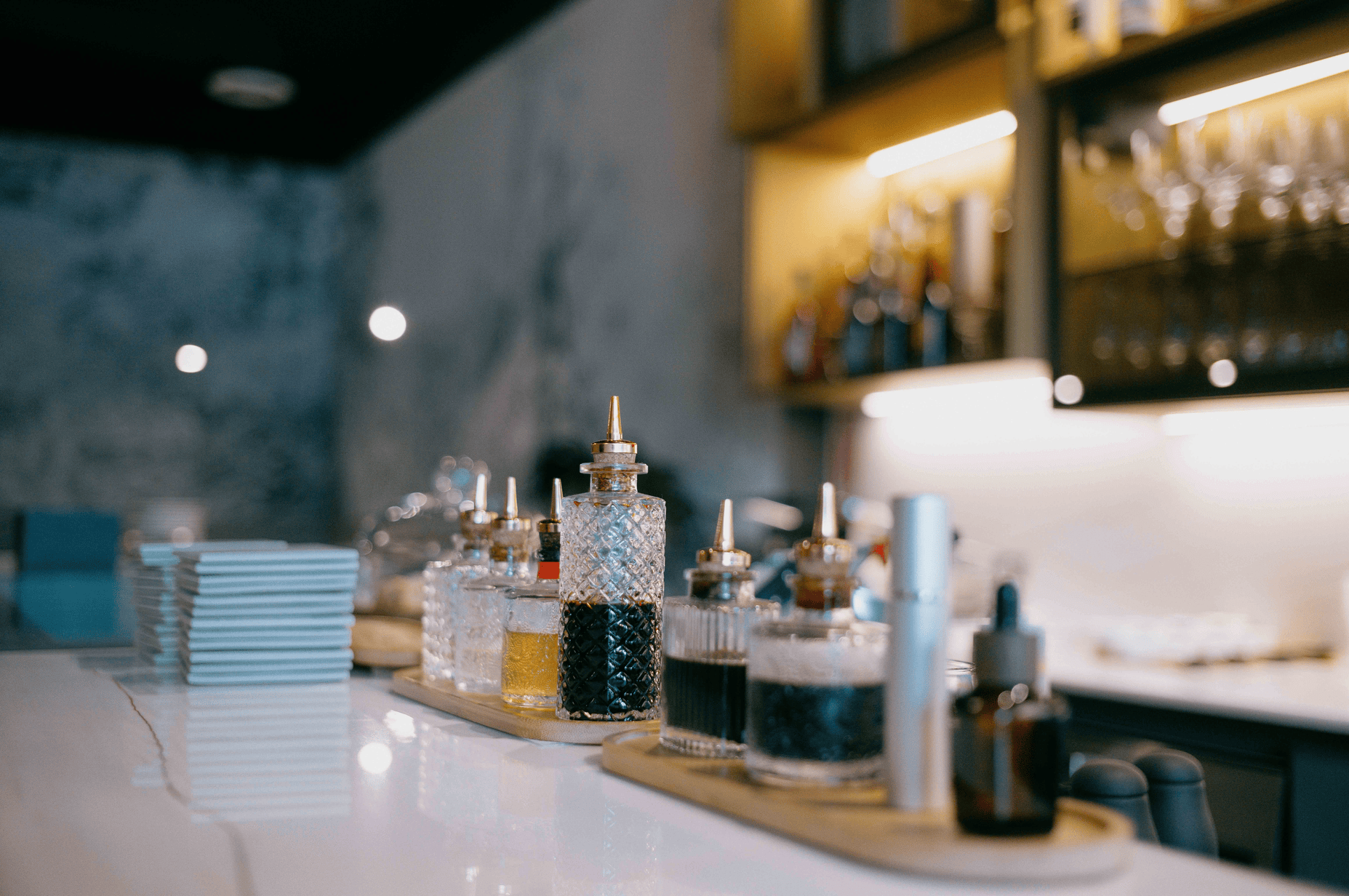 Glass bottles with pour spouts containing various liquids on a tray on a bar counter with stacked plates in the background.
