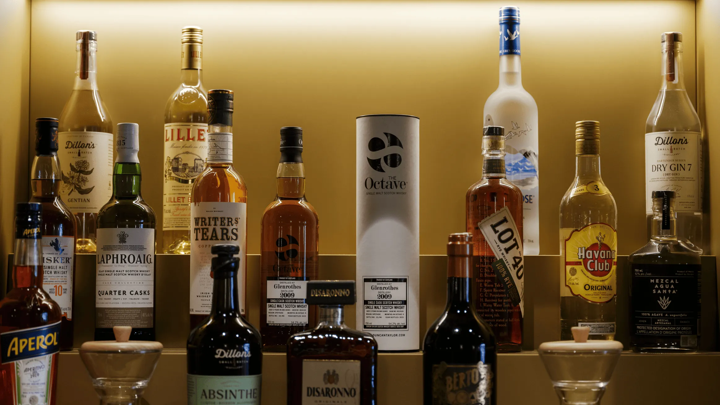 Shelf displaying various liquor bottles including Writer's Tears, Laphroaig, Havana Club, Grey Goose, and Disaronno under warm lighting.
