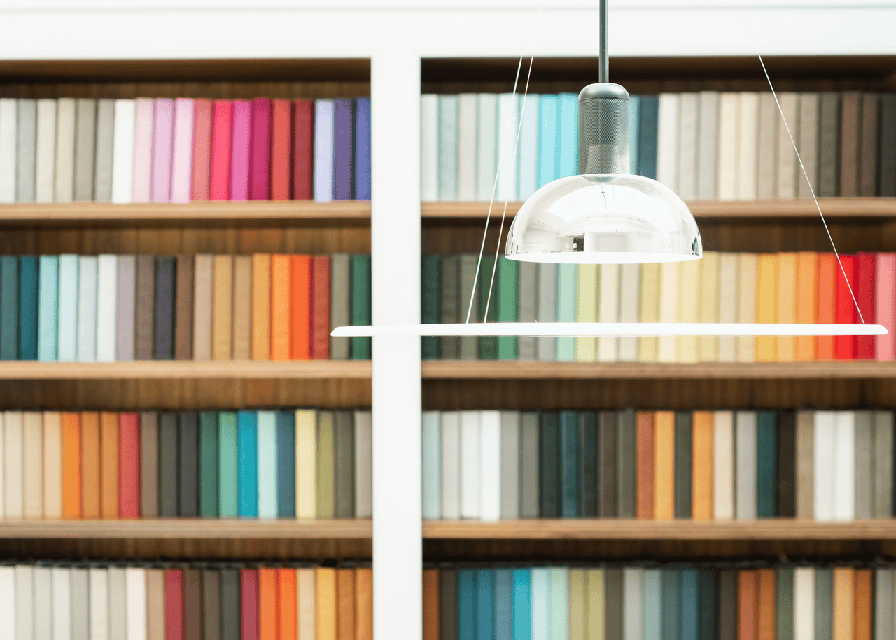 Modern silver pendant light hanging in front of shelves filled with colorful fabric swatches organized by color.