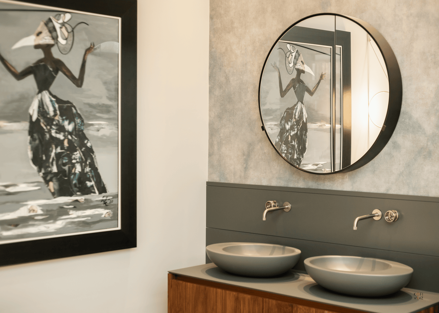 Modern bathroom with two round gray sinks, wall-mounted faucets, a round mirror, and abstract artwork reflected in the mirror.