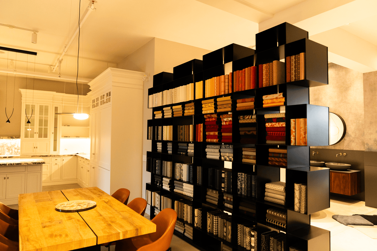 Bright modern interior with a wooden dining table and brown chairs next to a black shelving unit filled with neatly organized rolled fabrics in various colors.