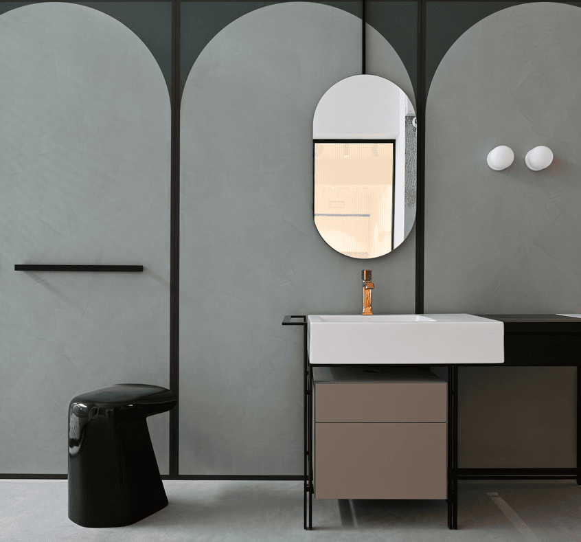 Modern bathroom with matte gray arched wall panels, an oval mirror, white rectangular sink with gold faucet, black stool, and minimalist black shelving.