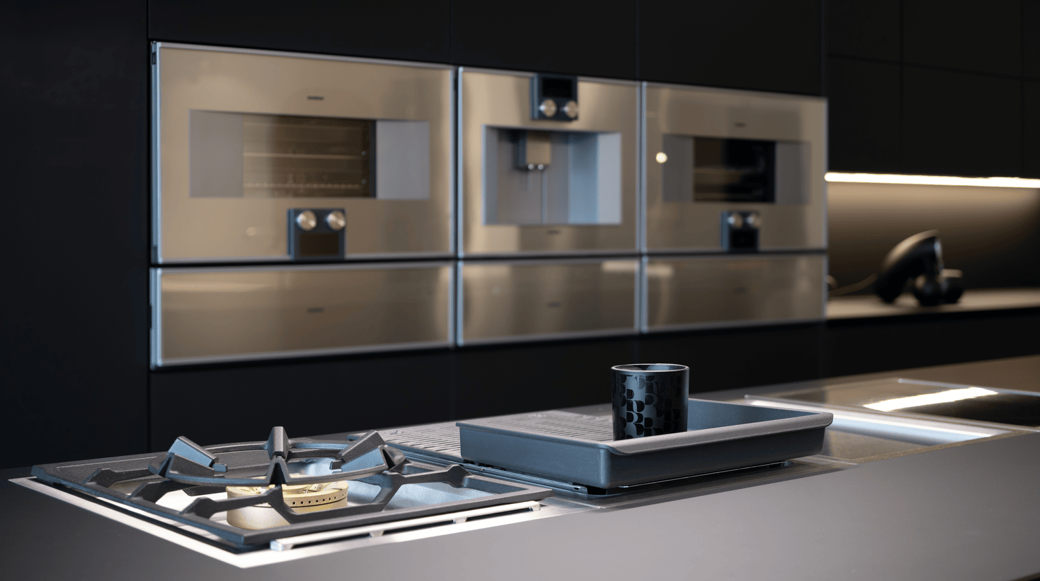Modern kitchen countertop with gas stove burner, baking tray, and a black patterned cup, sleek stainless steel built-in ovens in background.