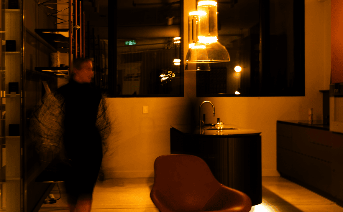 Blurry person walking through a dimly lit modern kitchen with warm lighting and a brown chair in the foreground.