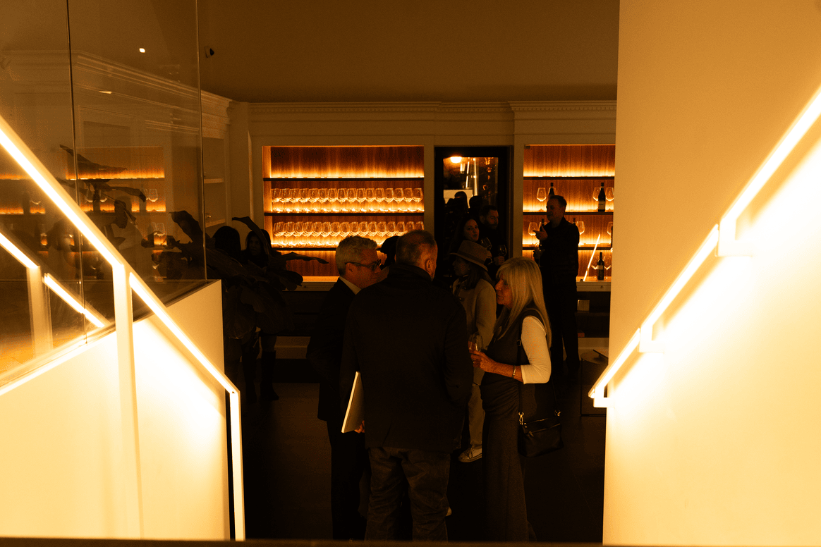 Dimly lit indoor social gathering with people conversing near illuminated shelves of glassware and bottles.