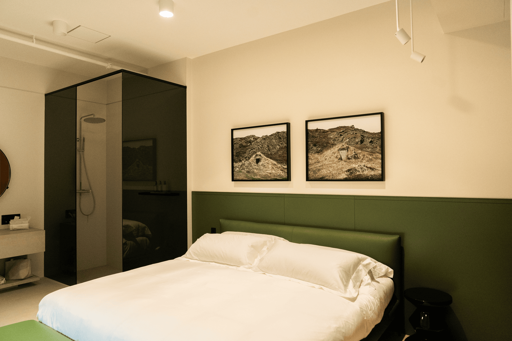 Modern bedroom with a large bed, green headboard, two landscape photos on the wall, and a glass-enclosed shower in the corner.