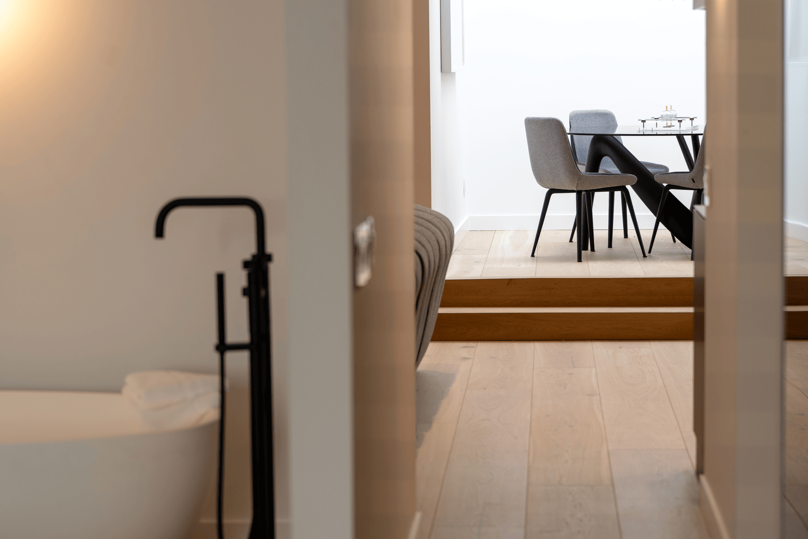 Minimalist interior showing a partial view of a freestanding white bathtub with a black floor-mounted faucet on the left and a dining area with gray chairs and a black table in the background on a wooden floor.