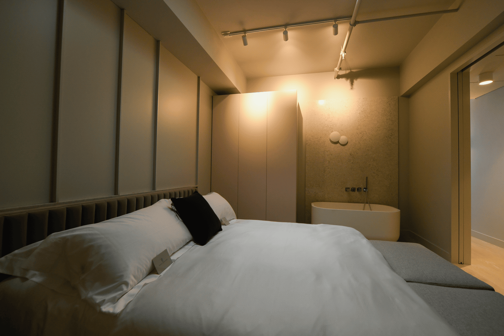 Modern bedroom with a white bed, black cushion, beige wardrobe, and a freestanding bathtub near a textured wall under warm lighting.