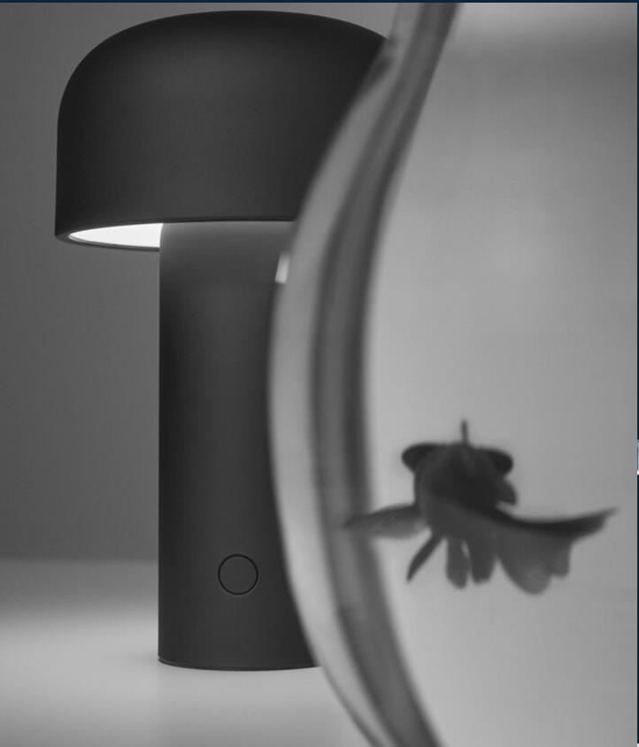 Black modern table lamp with a circular button on its base placed behind a partially visible fishbowl with a blurred goldfish inside.