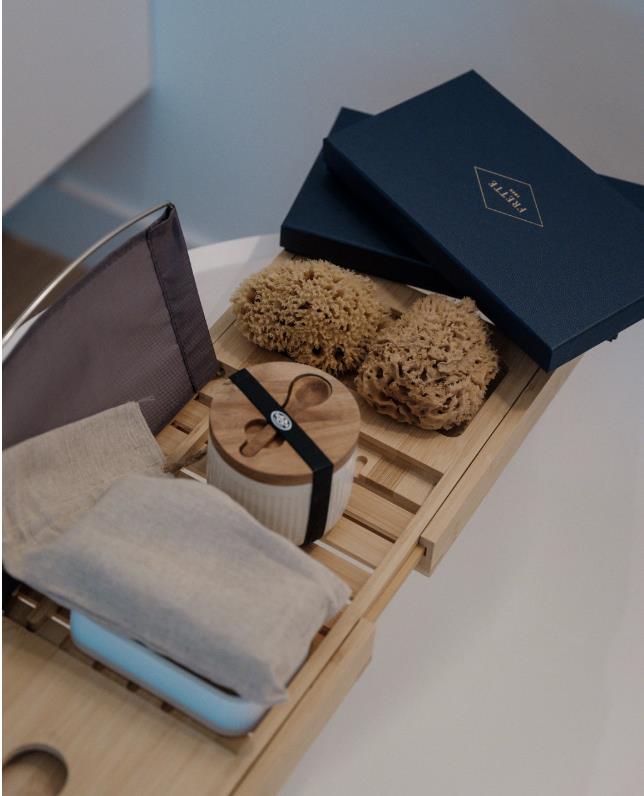 Wooden bath tray holding natural bath sponges, a ribbed white container with a wooden lid and spoon, a folded beige cloth, and two blue boxes.